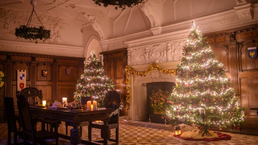 Two large Christmas trees with red and gold decorations and white fairy lights stand either side of a fireplace in a wood panelled room. A dark wooden table with candles is in the foreground.